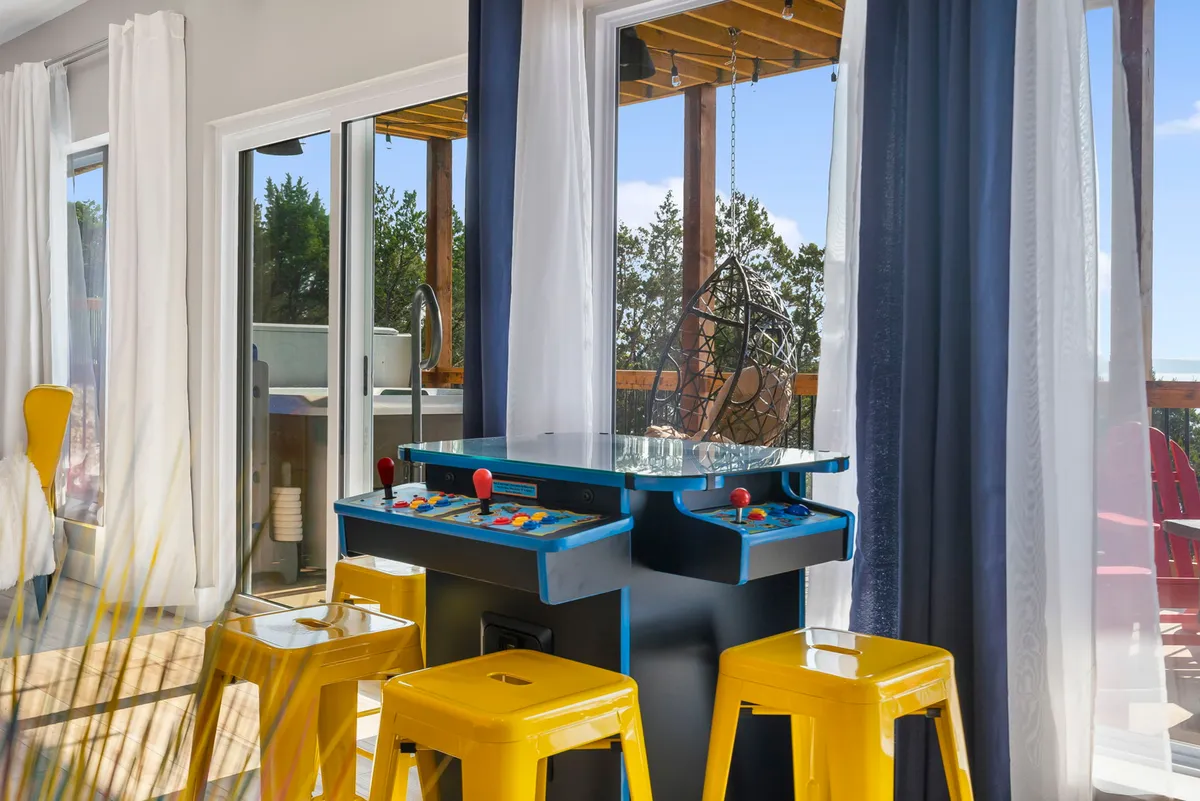 This image features an arcade game machine with bright yellow stools positioned in front of large windows, filling the space with natural light. The scene suggests an energetic, playful atmosphere where friends or family can gather for classic arcade fun.