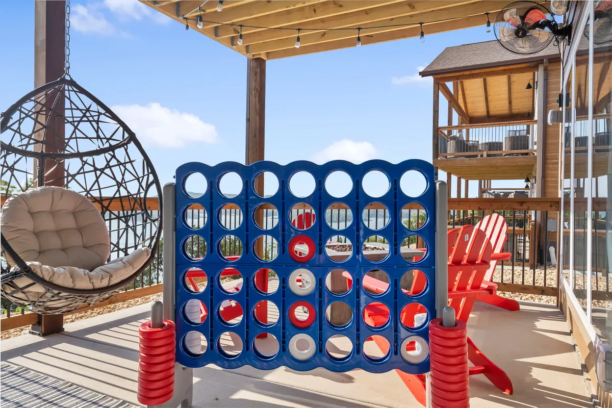 An exterior deck with a hanging chair, a life-sized Connect Four game set, and red Adirondack chairs overlooking a scenic, natural view. The space is designed for playful outdoor fun and gathering with friends or family.