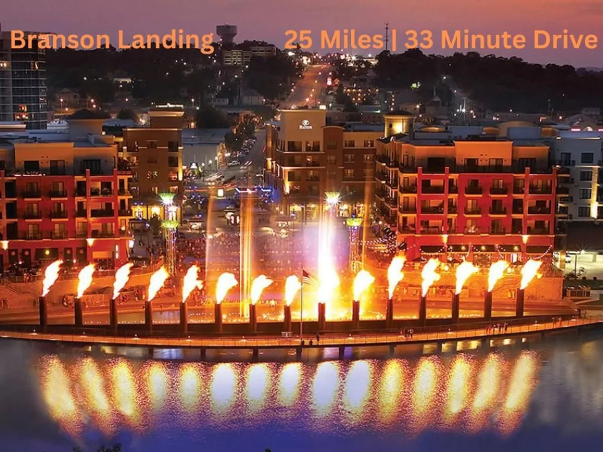 An evening waterfront scene at Branson Landing, highlighted by a spectacular fountain display, illuminated buildings, and shimmering reflections on the water. The lively atmosphere suggests dining, shopping, and entertainment options along the boardwalk.