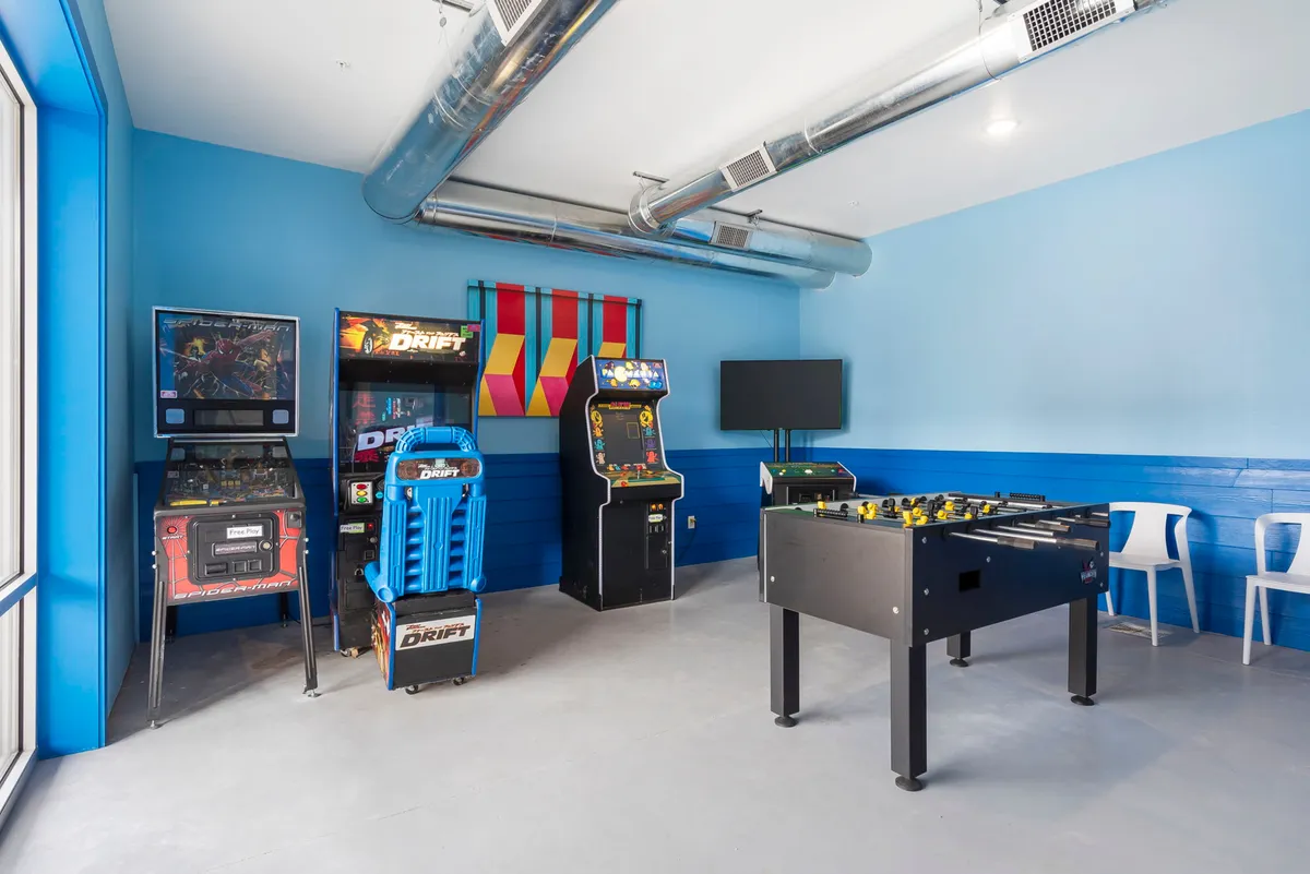A dynamic game room scene with multiple arcade machines, a foosball table, vibrant wall art, and a TV. The space feels energetic and bustling, ready for friendly competition and group gatherings.