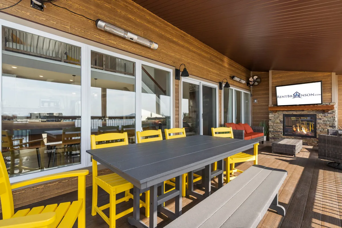 A cheerful deck scene featuring a dining table with yellow chairs, an outdoor fireplace, a television, and a lounge area with red cushions. The setup is perfect for alfresco dining, lively gatherings, and enjoying meals with a view.