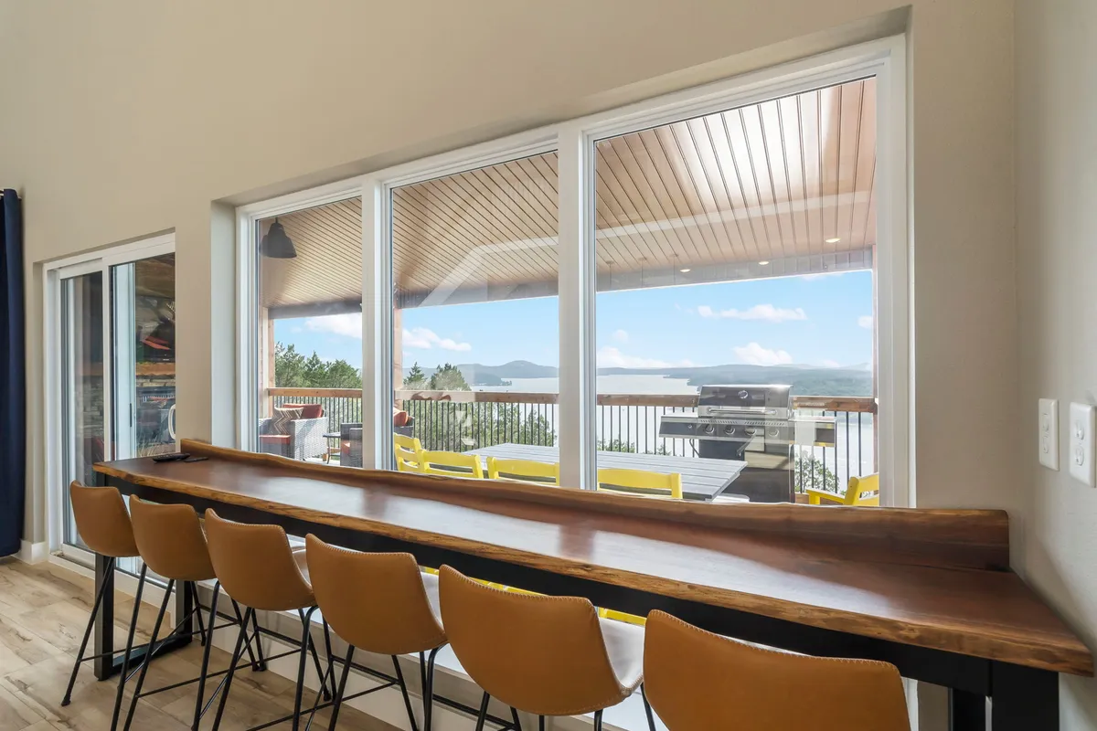 A bright, relaxing bar and dining area with floor-to-ceiling windows opening to an outdoor deck and gorgeous lake view. The custom wooden bar counter and stylish bar stools create an appealing spot for morning coffee or evening drinks while soaking in the scenery.