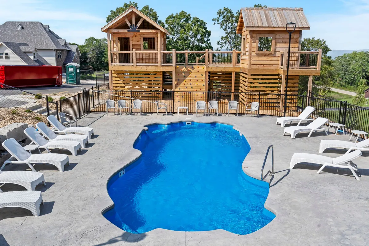 The image shows an inviting blue swimming pool surrounded by lounge chairs and wooden playhouses, with children playing and families relaxing nearby. The space is sun-drenched and vibrant, designed for summer fun, group entertainment, and memorable outdoor experiences for all ages.