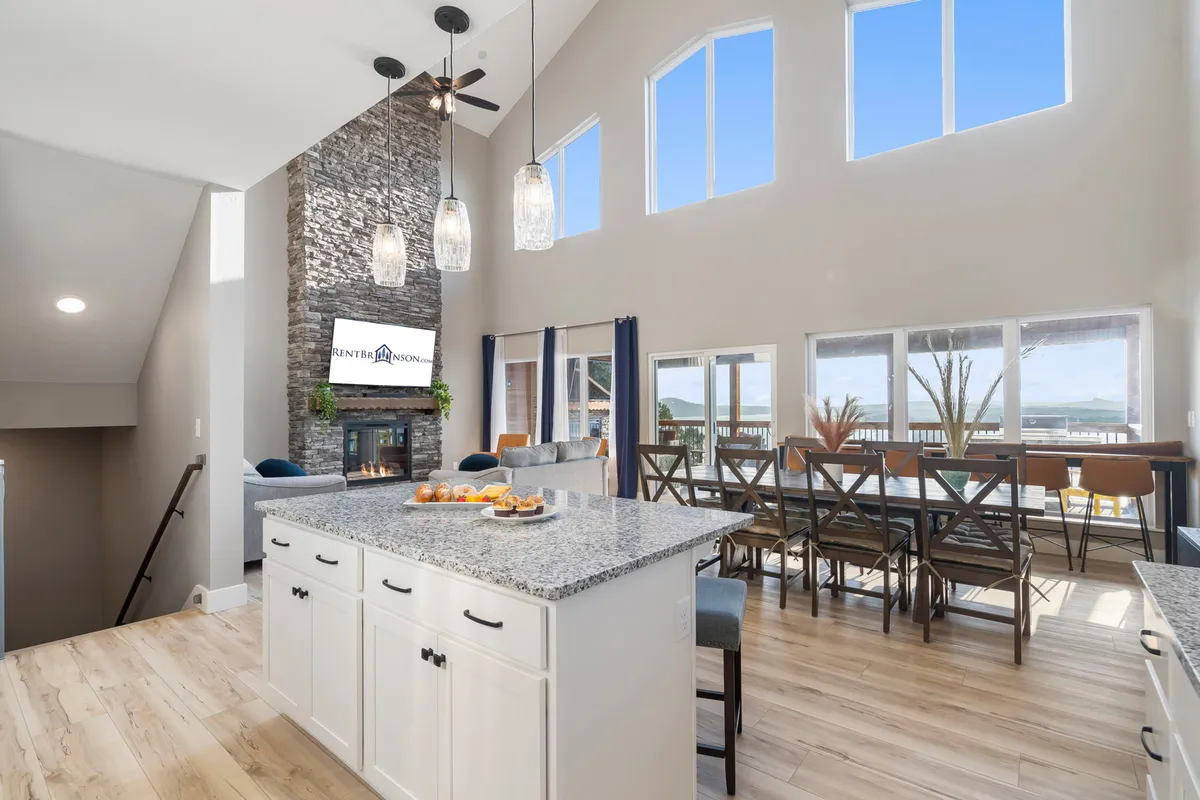 A welcoming communal space featuring a spacious kitchen island, stone fireplace, large windows framing the outdoors, and a stylish dining area. Pendant lighting and plush seating create an ideal setting for group meals, celebrations, or cozy evenings by the fire.