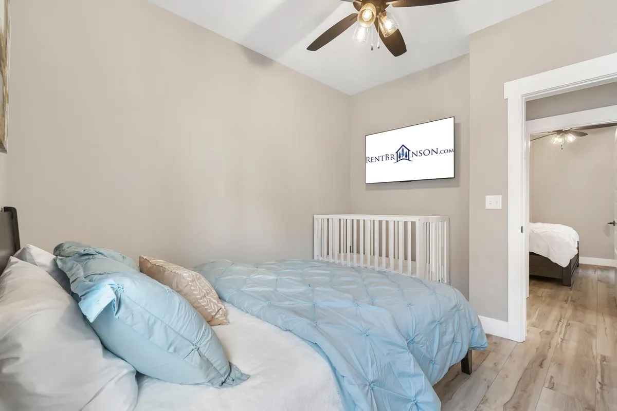 A serene bedroom with a bed covered in a blue quilt, a flat-screen TV, and a crib, blending restful adult space with baby-friendly amenities. Natural light filters in, creating a calming atmosphere suitable for families with young children.