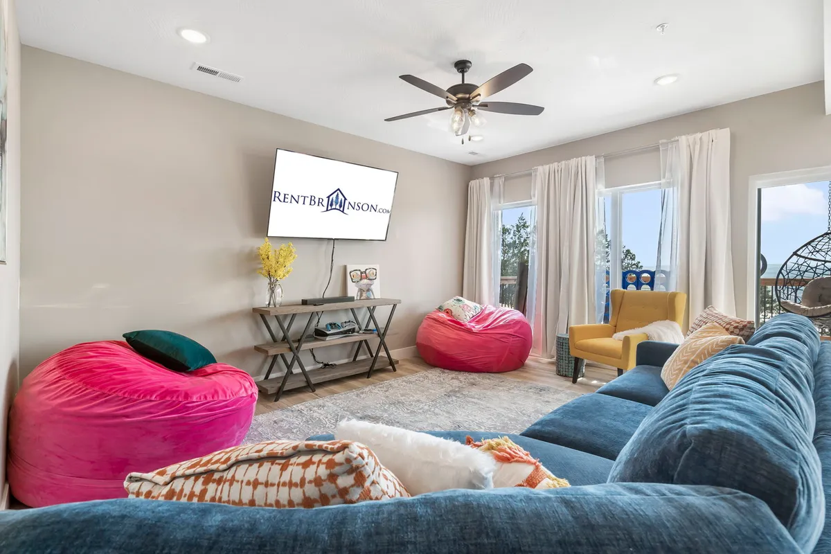 A spacious entertainment area featuring a large blue sectional sofa, bright pink bean bags, a television, and a pop of color from a yellow plant and a multicolored chair. The setup is perfect for movie marathons, board games, or chilling out after a day of adventure.
