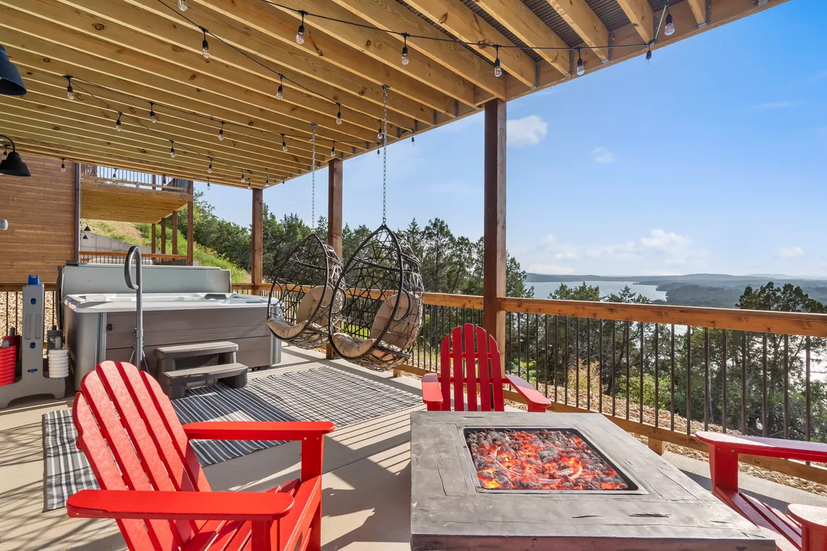 This exterior image features a spacious patio area equipped with hanging chairs, a fire pit surrounded by red Adirondack chairs, and a hot tub. The patio overlooks the lake, inviting guests to soak in the view while relaxing or socializing. Comfortable seating and leisure-focused amenities are front and center, exuding an atmosphere of relaxation and togetherness.