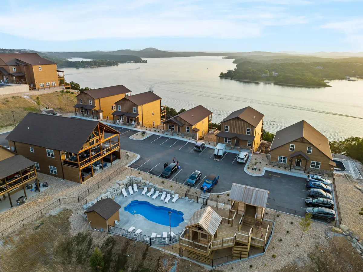 This exterior image captures a lively scene featuring a swimming pool with crystal-clear water, a nearby playground structure perfect for children, a scenic lake view in the background, and a row of cabins. The space is designed for multiple family-friendly activities, including swimming, gatherings, outdoor games, barbecuing, and relaxing by the lake.