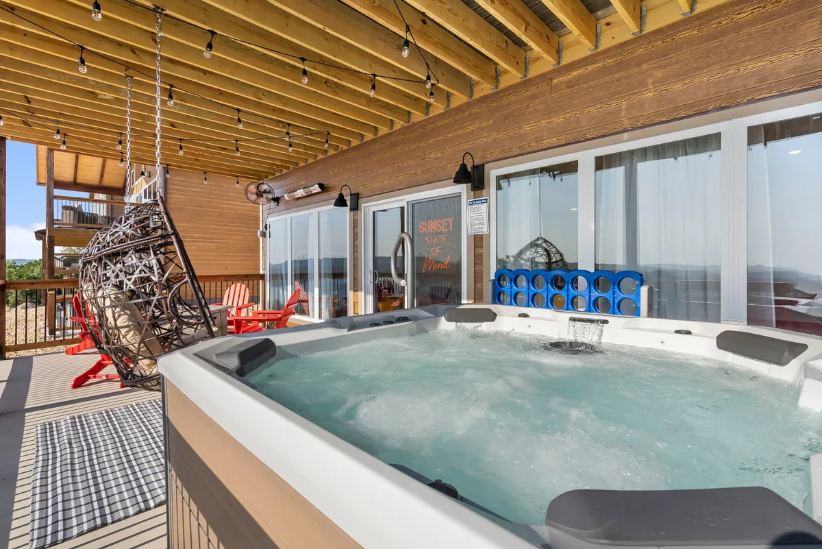 A tranquil patio area with a bubbling hot tub, hanging chair, red adirondack chairs, and stunning landscape views. The scene suggests relaxation and leisure, from soaking in the hot tub to reading in the hanging chair as the sun sets.