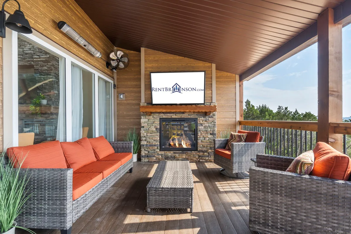 An inviting outdoor living area with a stone fireplace, comfortable seating, a mounted television, and lush greenery surrounding the deck. The space provides a cozy environment for relaxing by the fire or enjoying a game on the outdoor TV, with sunset views possible from the deck.