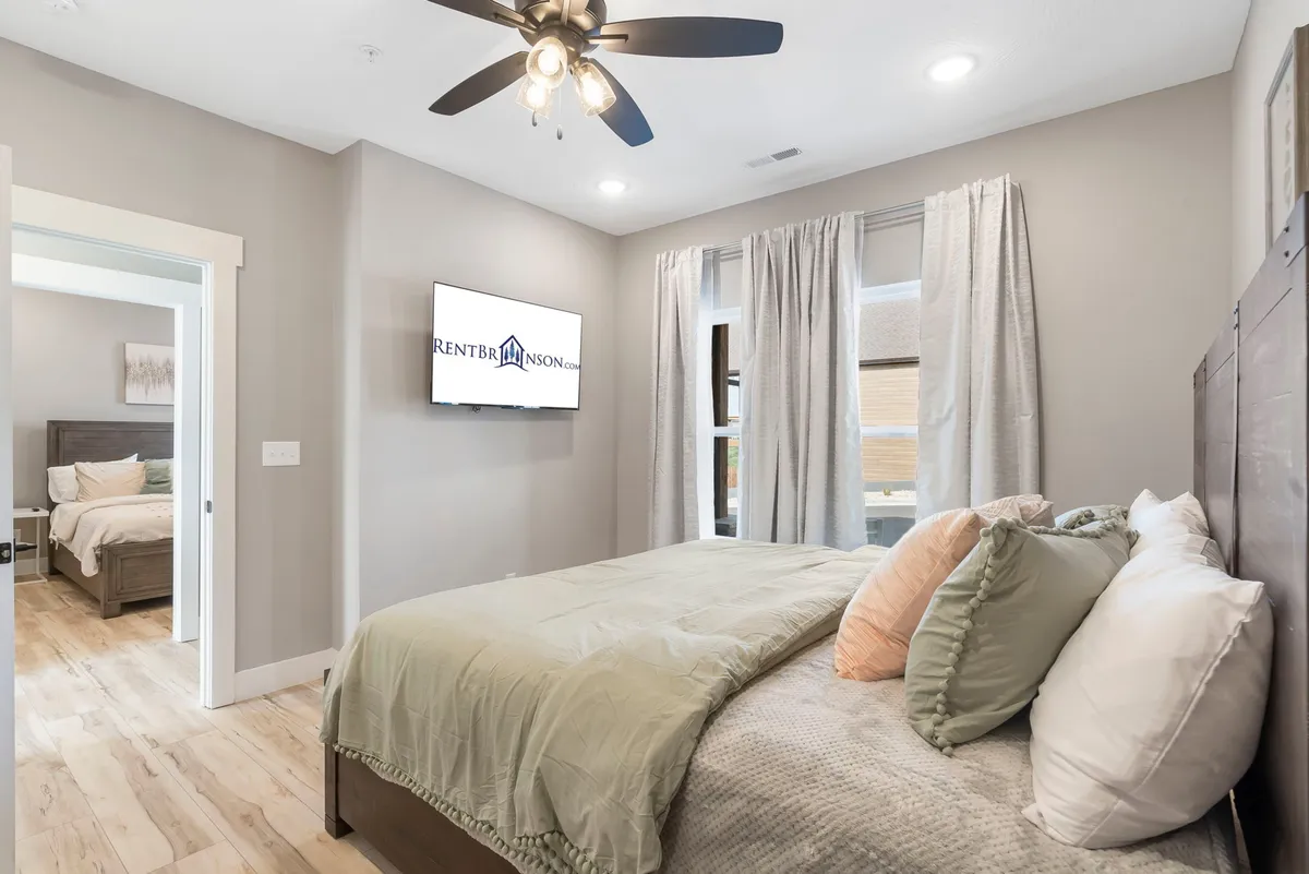A cozy bedroom with a plush bed adorned with decorative pillows, a TV mounted on the wall, and large windows with curtains that let in natural light. The setting exudes comfort and tranquility, ideal for relaxation and restful sleep.
